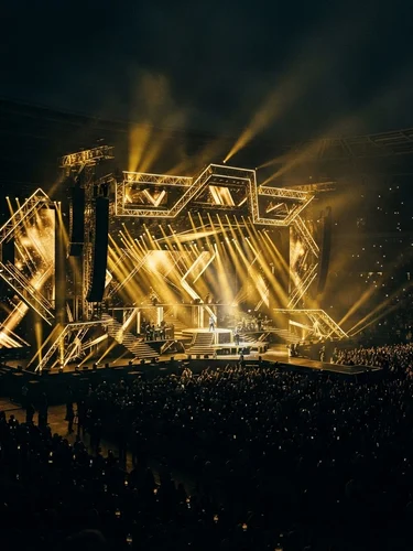 Dramatic wide-angle view of a monumental concert stage illuminated with gold architectural beam lights against a moody black stadium environment